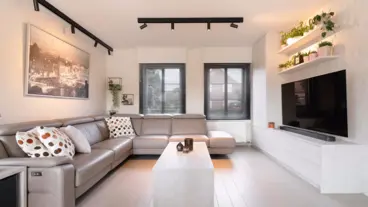 Modern woonkamer met lichtgrijze hoekbank, witte salontafel, zwarte jaloezieën en een grote tv aan de muur, versierd met groene planten.