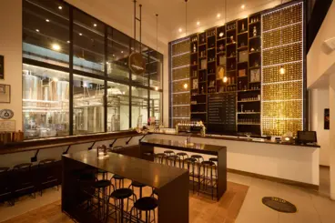Interior of a modern bar with a large black shelving unit displaying bottles and awards, wooden tables, and black bar stools, with a view of the brewing area through large glass panels.