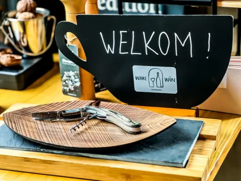 Een houten tafel met een houten snijplank en een kurkentrekker. Op de achtergrond een bord met verschillende koekjes en een bord met walnoten. Een zwart bord met de tekst 'Welkom!' en een logo met een glas en een fles wijn.
