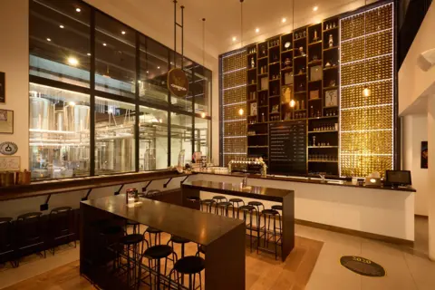 Interior of a modern bar with a large black shelving unit displaying bottles and awards, wooden tables, and black bar stools, with a view of the brewing area through large glass panels.