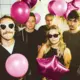 Vijf jongeren staan samen in een ruimte met witte tegels aan de muur, omringd door roze en paarse ballonnen, sommigen dragen zonnebrillen en feestkleding, en ze poseren voor een feest of feestje.