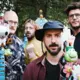 Vijf mannen poseert in een park met kleurrijke randobjecten in de hand. Ze kijken naar de camera, met achtergrond van groen bladerdak en een attractie die eruitziet als een draaimolen.