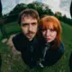 Tweedelige foto van een man en een vrouw die naar de camera kijken, genomen met een vislens. Beiden staan op een grasveld met bomen op de achtergrond, onder een bewolkte hemel.