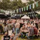 Een drukke enthousiaste menigte bij een outdoor festival met kleurrijke vlaggen en parasols, groene bomen op de achtergrond.