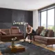 Vrouw zittend op een bruin leren hoekbank in een lichte woonkamer met grote ramen, zwarte muur en houten vloer.