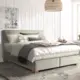Sfeervolle slaapkamer met een groot bed met neutrale kussens en dekens, houten bijzettafel met lamp, en een raam voor natuurlijk licht.