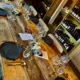 Een houten tafel is gedekt voor een wijnproeverij, met wijn- en waterglazen, borden, bestek en menu's. Op de achtergrond staan wijnflessen op houten planken.