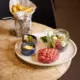 Raw ground beef patty, small jar of pickles, small jar of mustard, small bowl of mayonnaise, side salad with greens and vegetables, and a plate of French fries in a basket.