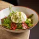 Salad with a poached egg, greens, cherry tomatoes, and green dressing in a white bowl with a brown rim on a stone table.