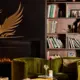 A modern cozy living room with a dark wall, a golden phoenix illustration, a fireplace, green velvet armchairs, a small round table with a flower in a glass jar, a magazine, and a bookshelf filled with books and decorative items.