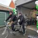 Twee mannen in zwarte kleding rijden op tandemfiets voor fietsenwinkel omringd door groene, witte en zwarte ballonnen
