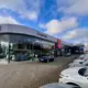 Moderne autodealer met Nissan en Suzuki merken, glazen gevel en geparkeerde auto's buiten onder een blauwe lucht met wolken